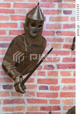 Medieval knight in chainmail stands against brick wall. Helmet with pointed top covers his face. He grips a sword with both hands. Brick wall behind him is textured and red Medieval knight in chainmail stands against brick wall. Helmet with pointed top covers his face. He grips a sword with both hands. Brick wall behind him is textured and red 141513143