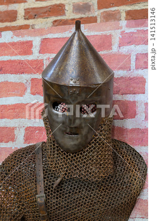Ancient metal helmet with chainmail armor. Red brick wall background. Helmet has eye slits and pointed top. Armor shows weathered, historical texture. Suitable for medieval or fantasy themes 141513146