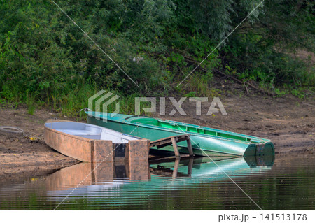 Green canoe rests beside wooden dock on calm water. Reflections shimmer softly in still lake surface. Lush green bushes line the muddy riverbank. Dusk light casts gentle hues on tranquil scene Green canoe rests beside wooden dock on calm water. Reflections shimmer softly in still lake surface. Lush green bushes line the muddy riverbank. Dusk light casts gentle hues on tranquil scene 141513178