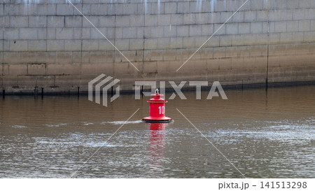 Red buoy floats calmly on water's surface. It marks a waterway or dock area clearly. Ripples gently surround the buoy's base. Background shows gray concrete wall edge Red buoy floats calmly on water's surface. It marks a waterway or dock area clearly. Ripples gently surround the buoy's base. Background shows gray concrete wall edge 141513298