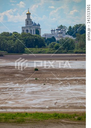 Bare riverbed shows muddy expanse and shallow waters. Ancient white church towers stand beyond green trees. Distant domes and bell tower grace the skyline Bare riverbed shows muddy expanse and shallow waters. Ancient white church towers stand beyond green trees. Distant domes and bell tower grace the skyline 141513301