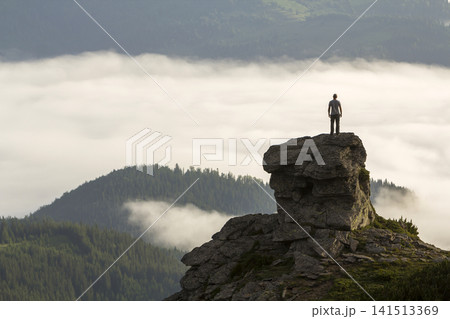 Silhouette of athletic climber tourist on high rocky formation on mountain valley filled with white puffy clouds and fog and covered with evergreen forest mountain slopes under clear sky background. Silhouette of athletic climber tourist on high rocky formation on mountain valley filled with white puffy clouds and fog and covered with evergreen forest mountain slopes under clear sky background. 141513369