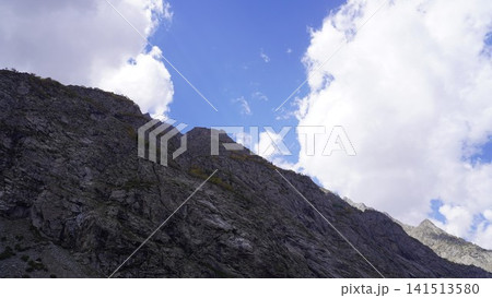 Dramatic photograph of a steep rocky mountain face against a bright blue sky and puffy clouds, imposing concept. 141513580