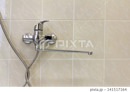 Water tap, chrome stainless metal faucet connected to shower stall tiled wall, mixer cold hot water on blurred bathroom copy space background. Modern home interior. Water tap, chrome stainless metal faucet connected to shower stall tiled wall, mixer cold hot water on blurred bathroom copy space background. Modern home interior. 141517164
