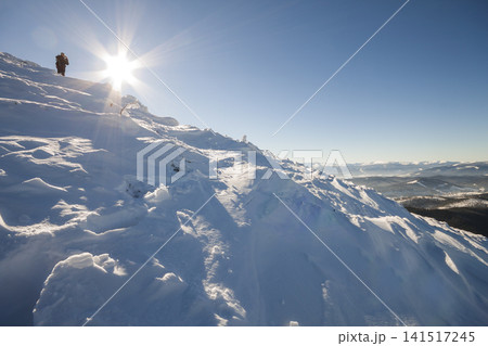Tourist hiker climber in winter clothing with backpack climbing dangerous rocky steep mountain slope covered with deep snow, white sun rays shining in bright blue sky copy space background. Tourist hiker climber in winter clothing with backpack climbing dangerous rocky steep mountain slope covered with deep snow, white sun rays shining in bright blue sky copy space background. 141517245