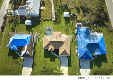 Top view of leaking house roof covered with protective tarp sheets against rain water leaks until replacement of asphalt shingles. Damage of building rooftop as aftermath of hurricane Ian in Florida Top view of leaking house roof covered with protective tarp sheets against rain water leaks until replacement of asphalt shingles. Damage of building rooftop as aftermath of hurricane Ian in Florida 141517250