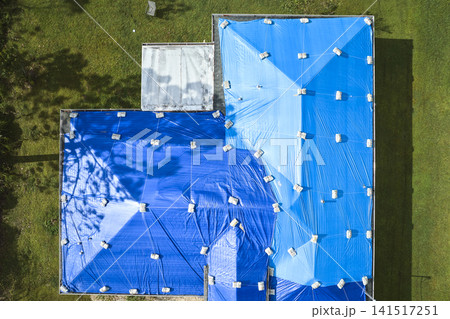 Top view of leaking house roof covered with protective tarp sheets against rain water leaks until replacement of asphalt shingles. Damage of building rooftop as aftermath of hurricane Ian in Florida Top view of leaking house roof covered with protective tarp sheets against rain water leaks until replacement of asphalt shingles. Damage of building rooftop as aftermath of hurricane Ian in Florida 141517251