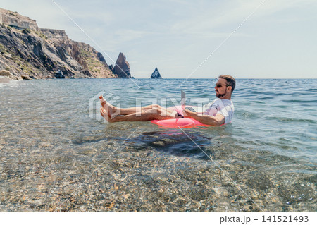 Freelancer laptop ocean man working remotely while floating on an inflatable ring in clear tropical waters near rocky cliffs Freelancer laptop ocean man working remotely while floating on an inflatable ring in clear tropical waters near rocky cliffs 141521493