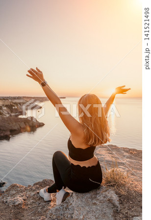 Sunset woman cliff sitting on a rocky ledge with arms raised toward the sun over the ocean reflecting peace and freedom Sunset woman cliff sitting on a rocky ledge with arms raised toward the sun over the ocean reflecting peace and freedom 141521498