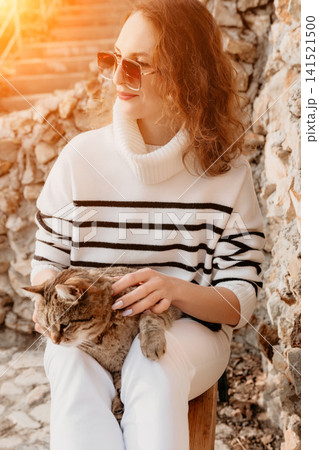 Woman cat pet sitting outside in striped sweater and sunglasses petting a tabby on her lap in warm golden sunlight 141521500