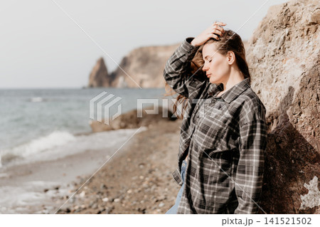 Woman beach cliffs young pensive woman in plaid shirt leaning against a boulder on a stony beach with distant cliffs Woman beach cliffs young pensive woman in plaid shirt leaning against a boulder on a stony beach with distant cliffs 141521502