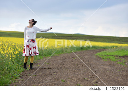 Woman hitchhiking on a dirt road next to a yellow canola flower field Woman hitchhiking on a dirt road next to a yellow canola flower field 141522191