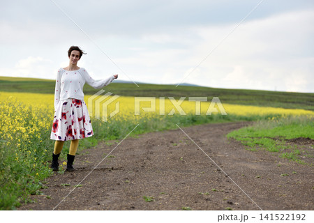 Woman hitchhiking on a dirt road next to a yellow canola flower field Woman hitchhiking on a dirt road next to a yellow canola flower field 141522192