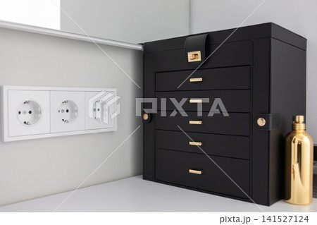 Black wooden jewelry organizer with small drawers stands on a light shelf beside decorative figurines and wall outlets, showing elegant home storage and modern interior decor view Black wooden jewelry organizer with small drawers stands on a light shelf beside decorative figurines and wall outlets, showing elegant home storage and modern interior decor view 141527124