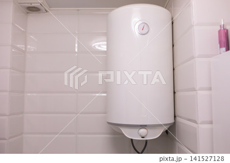 White electric water heater mounted on a tiled bathroom wall, showing a compact household appliance for hot water supply, modern plumbing, utility equipment and home comfort use 141527128