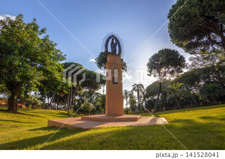 Religious statue of Virgin Mary on tall brick pedestal in green park surrounded by pine trees at La Rabida in Huelva Andalusia Spain under sunny blue sky. Religious statue of Virgin Mary on tall brick pedestal in green park surrounded by pine trees at La Rabida in Huelva Andalusia Spain under sunny blue sky. 141528041