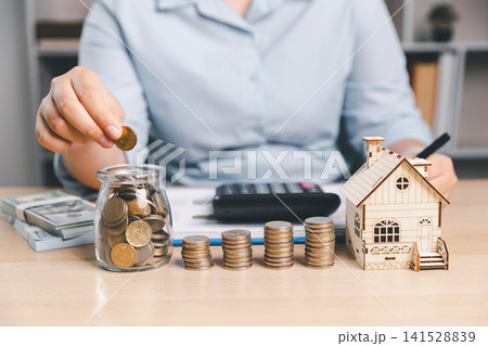 Person saving coins in glass money jar with wooden house model, calculator, cash and coin stacks on table, representing mortgage savings and real estate financial planning. Person saving coins in glass money jar with wooden house model, calculator, cash and coin stacks on table, representing mortgage savings and real estate financial planning. 141528839