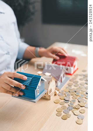 Property investment portfolio concept with hand selecting blue house model among homes and coins, showing real estate choice, home buying budget, mortgage planning, asset value, and wealth strategy. Property investment portfolio concept with hand selecting blue house model among homes and coins, showing real estate choice, home buying budget, mortgage planning, asset value, and wealth strategy. 141528914