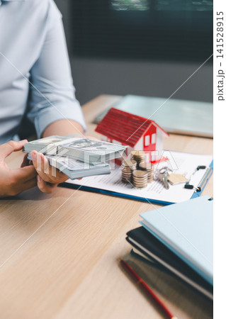 Property purchase payment concept with hands holding cash beside red house model, coin stacks, keys, and contract documents for mortgage deposit, home buying, finance, and real estate deal. 141528915