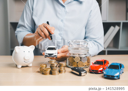 Business person planning car purchase savings with piggy bank, coin jar, toy cars, car key, cash, and coin stacks, representing auto loan, vehicle finance, and ownership budget. Business person planning car purchase savings with piggy bank, coin jar, toy cars, car key, cash, and coin stacks, representing auto loan, vehicle finance, and ownership budget. 141528995