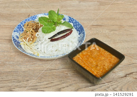 rice noodles topping fried chili and fresh vegetable on plate with sweet coconut milk mashed bean curry sauce in bowl 141529291