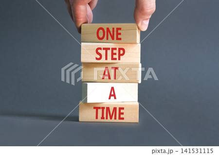 One step at a time symbol. Concept words One step at a time on beautiful wooden blocks. Beautiful grey background. Businessman hand. Business and one step at a time concept. Copy space. One step at a time symbol. Concept words One step at a time on beautiful wooden blocks. Beautiful grey background. Businessman hand. Business and one step at a time concept. Copy space. 141531115