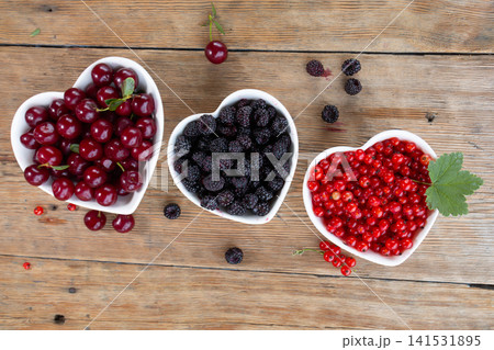 Three heart shaped white bowls filled with cherries blackberries and redcurrants on rustic wooden tabletop, viewed from above with scattered berries, fresh leaves and vibrant color Three heart shaped white bowls filled with cherries blackberries and redcurrants on rustic wooden tabletop, viewed from above with scattered berries, fresh leaves and vibrant color 141531895