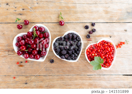 Three heart-shaped bowls filled with cherries, blackberries and red currants on rustic wooden table, top view with scattered berries, green leaf accent and bright natural food styling Three heart-shaped bowls filled with cherries, blackberries and red currants on rustic wooden table, top view with scattered berries, green leaf accent and bright natural food styling 141531899