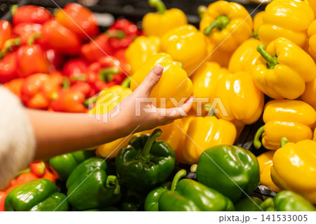 Woman hand choosing fresh yellow bell pepper in supermarket produce section, colorful vegetables displayed for healthy grocery shopping 141533005