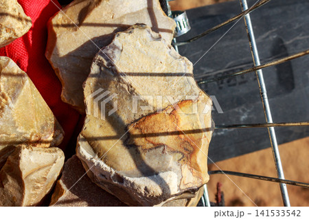 Sharp-edged flint stone shaped like a prehistoric hand axe, showing rough texture and ancient craftsmanship, set against a dark metallic grid background in bright sunlight Sharp-edged flint stone shaped like a prehistoric hand axe, showing rough texture and ancient craftsmanship, set against a dark metallic grid background in bright sunlight 141533542