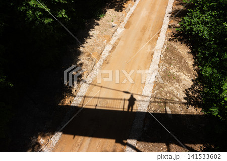 High-angle view of a dusty dirt path beneath a pedestrian bridge casting a dark silhouette of a lone person on the rural road surrounded by lush green summer forest foliage 141533602