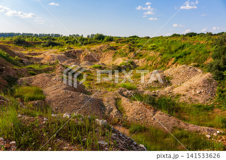 Overgrown rustic stone quarry with sandy mounds, reclaimed by nature, featuring patchy grass and wild vegetation under a bright summer sky in a rural countryside setting Overgrown rustic stone quarry with sandy mounds, reclaimed by nature, featuring patchy grass and wild vegetation under a bright summer sky in a rural countryside setting 141533626