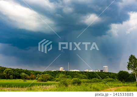 Dramatic dark storm clouds looming over a green forest landscape with city buildings in the distance, capturing an intense moody atmosphere before heavy rain falls on the horizon Dramatic dark storm clouds looming over a green forest landscape with city buildings in the distance, capturing an intense moody atmosphere before heavy rain falls on the horizon 141533885