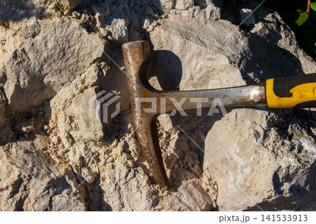 A weathered geological hammer rests against a textured limestone rock face in a quarry, capturing the essence of scientific field exploration and stone mineral research under bright sunlight A weathered geological hammer rests against a textured limestone rock face in a quarry, capturing the essence of scientific field exploration and stone mineral research under bright sunlight 141533913