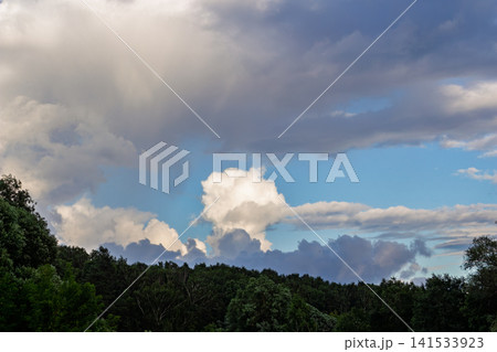 Dramatic massive white cumulus clouds drifting across a bright blue sky above a lush green forest canopy, capturing the vibrant atmosphere of a peaceful summer afternoon in nature Dramatic massive white cumulus clouds drifting across a bright blue sky above a lush green forest canopy, capturing the vibrant atmosphere of a peaceful summer afternoon in nature 141533923
