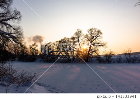 Golden hour paints a serene winter river landscape. Sun's vibrant light streams through bare trees, casting long shadows across the pristine snow. Pure natural beauty Golden hour paints a serene winter river landscape. Sun's vibrant light streams through bare trees, casting long shadows across the pristine snow. Pure natural beauty 141533941
