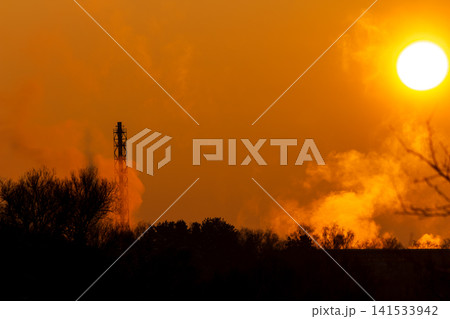 Golden winter sunset illuminates a towering industrial smokestack, its warm steam rising against the vibrant sky. Silhouetted trees below complete the dramatic scene 141533942
