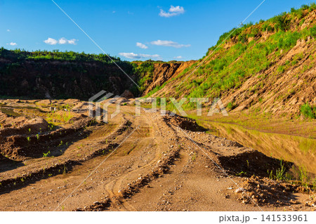 Abandoned sand quarry with muddy puddles, tire tracks, and green grass patches on steep slopes under a bright blue sky, showing post-mining natural reclamation in the countryside Abandoned sand quarry with muddy puddles, tire tracks, and green grass patches on steep slopes under a bright blue sky, showing post-mining natural reclamation in the countryside 141533961
