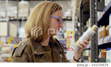 Young woman carefully choosing cosmetics in supermarket reading label on bottle, making informed purchasing decisions about health and beauty products Young woman carefully choosing cosmetics in supermarket reading label on bottle, making informed purchasing decisions about health and beauty products 141534674
