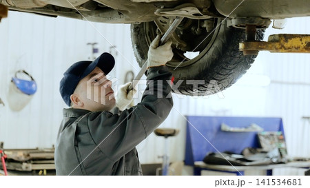 Professional mechanic leveraging specialized tool while repairing car suspension, working inside automotive garage with lifted vehicle Professional mechanic leveraging specialized tool while repairing car suspension, working inside automotive garage with lifted vehicle 141534681