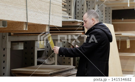 Skilled carpenter comparing lumber prices on smartphone, holding particle board sample in hardware store warehouse, evaluating material options for construction project 141534682
