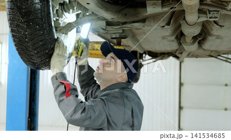 Mechanic wearing protective gear inspecting car undercarriage using flashlight on hydraulic lift inside professional automotive workshop Mechanic wearing protective gear inspecting car undercarriage using flashlight on hydraulic lift inside professional automotive workshop 141534685
