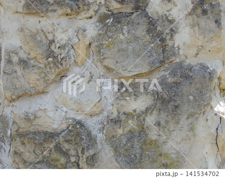 An old stone wall made of uneven beige and gray cobblestones. The rough, textured surface reveals traces of light-colored cement mortar, a dark layer of weathering, and patches of moss. An old stone wall made of uneven beige and gray cobblestones. The rough, textured surface reveals traces of light-colored cement mortar, a dark layer of weathering, and patches of moss. 141534702