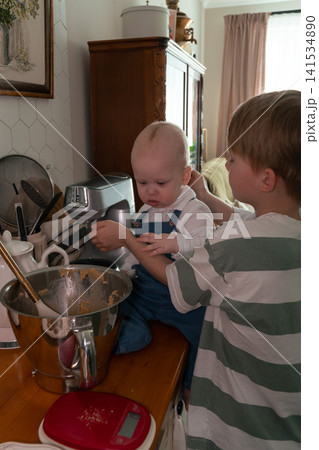 Older brother holds baby sibling beside bowl of homemade dough in family kitchen during baking preparation at home. Family, sibling bond, parenting, childhood, home cooking, lifestyle, togetherness Older brother holds baby sibling beside bowl of homemade dough in family kitchen during baking preparation at home. Family, sibling bond, parenting, childhood, home cooking, lifestyle, togetherness 141534890