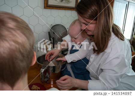 Mother helps baby stir homemade dough while older child joins family baking activity in cozy kitchen at home. Family, parenting, childhood, home cooking, bonding, education, lifestyle, togetherness Mother helps baby stir homemade dough while older child joins family baking activity in cozy kitchen at home. Family, parenting, childhood, home cooking, bonding, education, lifestyle, togetherness 141534897