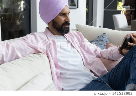 Indian man wearing turban holding smartphone while sitting on sofa in living room 141538992