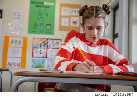 Female child student writing in notebook with yellow pencil at school desk next to window 141538994