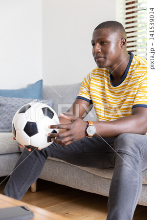 African american man sitting on sofa holding soccer ball in living room with tablet 141539014