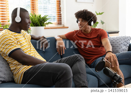 Diverse male friends recording podcast on blue sofa in living room with microphone, headphones 141539015