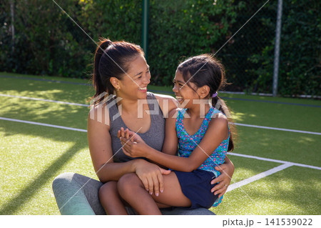 Mom and daughter smiling and sitting on turf court with white lines chain-link fence hedge Mom and daughter smiling and sitting on turf court with white lines chain-link fence hedge 141539022